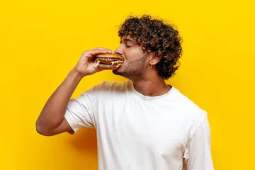 Schilderijen op glas young hungry indian guy eating delicious cheeseburger on yellow isolated background, curly man eating fast food and biting burger © Богдан Маліцький