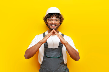 young indian male builder in hard hat and overalls showing house roof and protection on yellow...