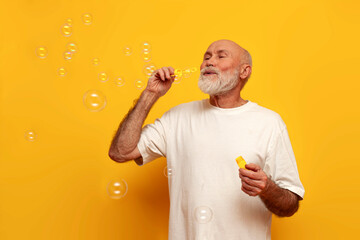 cheerful old bald grandfather in white t-shirt blowing soap bubbles on yellow isolated background,...