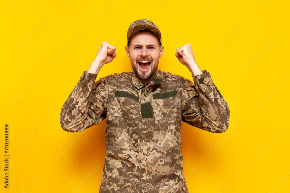 Sticker young Ukrainian male soldier in pixel camouflage uniform celebrates victory on a yellow isolated background, guy military cadet of the Ukrainian army wins and rejoices at luck and success