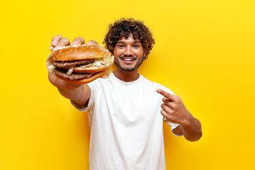 young curly indian man showing a big tasty burger and pointing with his hand on a yellow isolated background, the guy advertises fast food and recommends a cheeseburger and gives a hamburger - obrazy, fototapety, plakaty