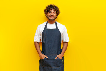 young Indian male barista in an apron stands on a yellow isolated background, a Hindu guy waiter in...