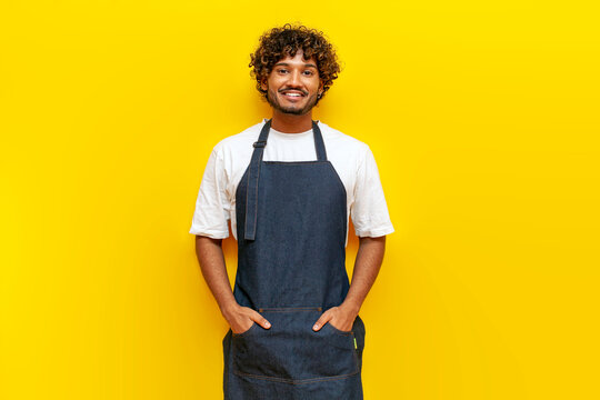 young Indian male barista in an apron stands on a yellow isolated background, a Hindu guy waiter in uniform smiles and looks at the camera