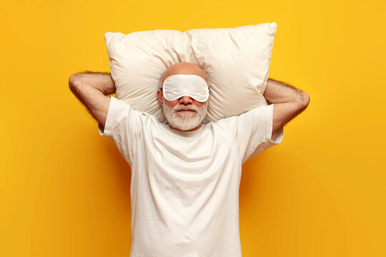 cheerful old bald grandfather in white sleep mask lies on soft comfortable pillow and rests on yellow isolated background, elderly pensioner is ready for bed relaxing and smiling