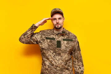 young Ukrainian male soldier in pixel camouflage uniform salutes on a yellow isolated background,...