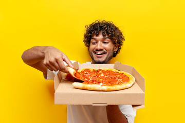 young hungry indian guy holding a box of delicious pizza and taking a piece on a yellow isolated...