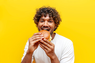 young hungry indian guy eating delicious cheeseburger on yellow isolated background, curly man...