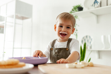 funny little boy 2 years old eats breakfast himself in the kitchen with a dairy product, the child...