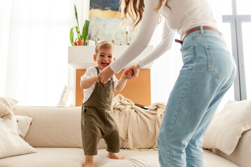 young mother and her little son dance and jump on the sofa at home, 2-year-old boy screams and rejoices with his parent, woman has fun with her child