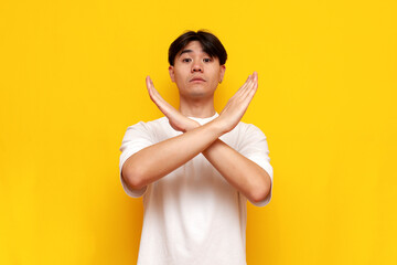 asian man in white t-shirt shows stop and prohibit gesture with crossed arms on yellow isolated...