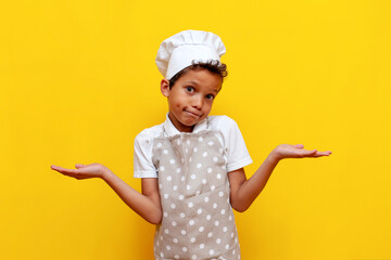 unsure african american boy in uniform and chef's hat shrugs his shoulders on yellow isolated background, confused teenager child in apron raises his hands and shows uncertainty