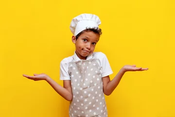 Fotobehang unsure african american boy in uniform and chef's hat shrugs his shoulders on yellow isolated background, confused teenager child in apron raises his hands and shows uncertainty © Богдан Маліцький