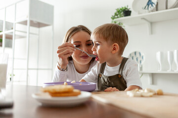 young mother spoon-feeds her little son and smiles, 2-year-old boy has breakfast in the kitchen with his parent, woman gives food to the child