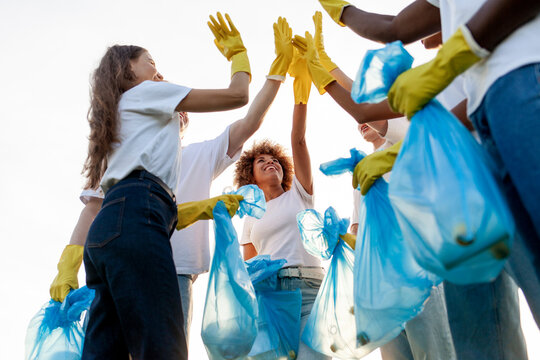 team of volunteers in gloves and with garbage bags rejoice together at success and high five, group of multiracial people hold hands and clean the park from garbage and plastic