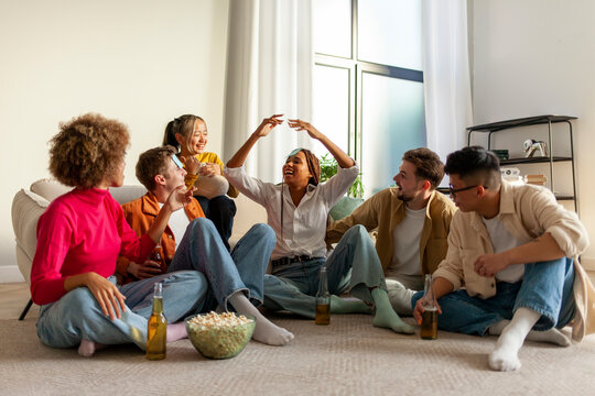 multiracial group of young people sitting at home with beer and popcorn and playing charades and having fun with friends, students of different ethnicities at home playing games and celebrating