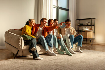 multiracial group of young friends sitting on sofa at home hugging and looking at camera, group of students watching TV together in room and smiling