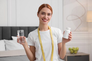 Happy young woman with bottle of pills, water and measuring tape at home. Weight loss