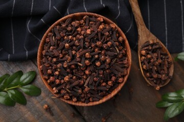 Aromatic cloves in bowl, spoon and green leaves on wooden table, flat lay