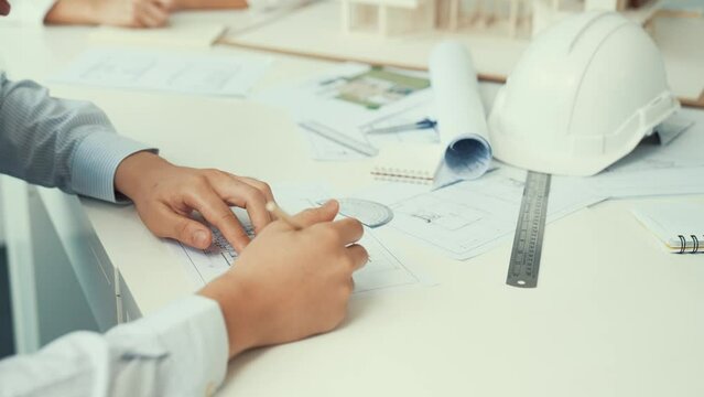 Close up of skilled architect engineer hand draws blueprint on meeting table with blueprint, architectural equipment, house model and safety helmet scatter around. Focus on hand. Immaculate.