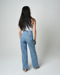 Full length portrait of brunette female model wearing casual clothes, white singlet shirt, denim jean pants. Standing pose walking away from camera. Isolated on white studio background.