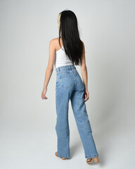 Full length portrait of brunette female model wearing casual clothes, white singlet shirt, denim jean pants. Standing pose walking away from camera. Isolated on white studio background.