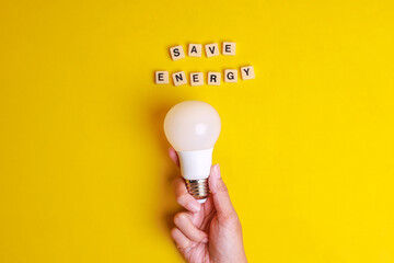 Save Energy words written on wooden blocks with hand holding white light bulb on yellow background. Concept of saving energy by using LED lightbulb - obrazy, fototapety, plakaty