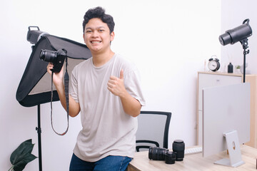 Young photographer smiling while holding camera and giving a thumb up at office studio.