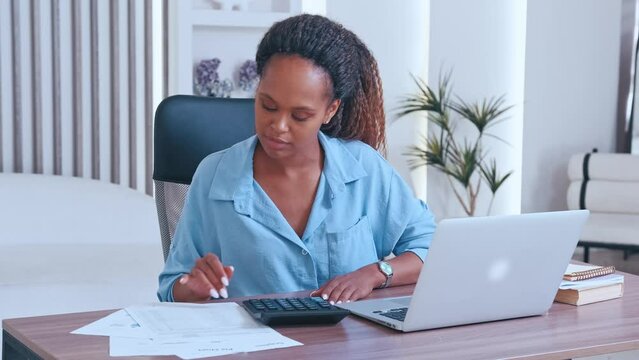Young focused smart African American woman calculates taxes by filling out online declaration and checking with paper documents to avoid mistakes sits at office desk in office. Businesswoman lifestyle