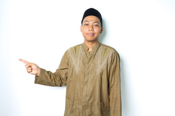 calm asian muslim man smiling at camera wearing koko clothes pointing to beside isolated on white...