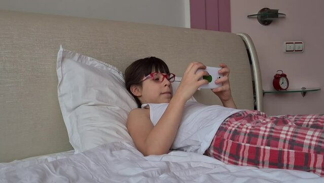 Teenage child with poor vision wearing glasses squints and looks at screen lying in bed. Farsightedness nearsightedness astigmatism glaucoma and gadget addiction