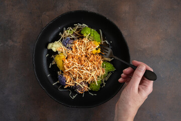 Bowl of cooked rainbow cauliflower topped with shredded chicken and grated parmesan cheese with a fork held by a woman’s hand, a nutritious meal

