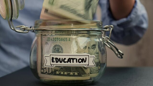 Unrecognizable woman take money from Saving Money In Glass Jar filled with Dollars banknotes. EDUCATION transcription in front of jar. Managing personal finances extra income for future insecurity