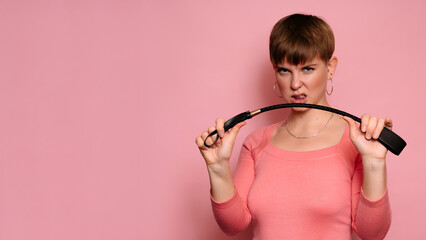A photo portrait of a young woman with a leather whip in her hands, highlighted on a bright pink...