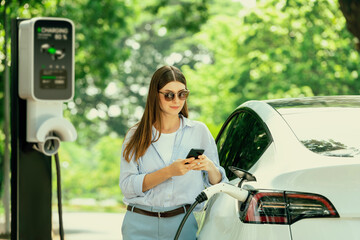 Young woman using smartphone online banking application to pay for electric car battery charging...
