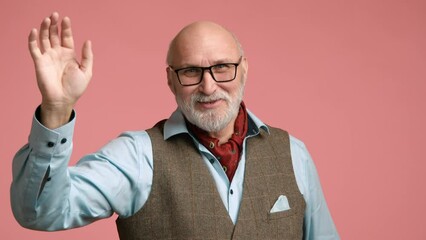 A senior man in a blue shirt and tweed vest is greeting with a raised hand, showcasing a friendly demeanor on a pink background. Camera 8K RAW. 