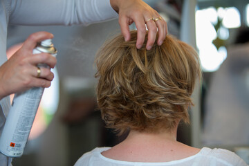 Fototapeta premium The image captures a moment of hairstyling where hands gently sculpt a short, layered haircut while applying hairspray to set the style in place. The hairstylist's hands are adorned with a wedding