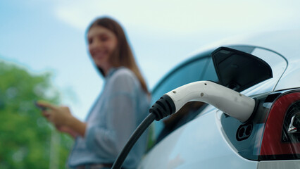 Young woman recharging battery for electric car during road trip travel EV car in natural forest or...