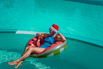 A happy woman in a blue bikini, a red and white Santa hat and sunglasses poses in the pool in an inflatable circle with a watermelon pattern, holding a glass of champagne in her hands.