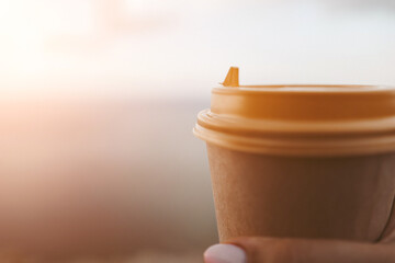 Hand holding Yellow cup with lid, coffee against a backdrop of a blue sky and sea. Illustrating cup...