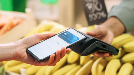 Close Up of a Customer Making a Secure Swift Contactless Payment Using a Smartphone with a Credit Card Banking NFC App. Modern Shopper Buying Organic Fruits and Vegetables From a Farm Market Stall
