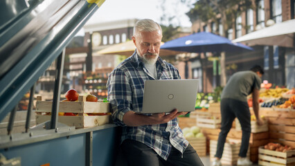 Middle Aged Businessman Working on Laptop Computer, Contacting Farm Suppliers About Fresh Produce for a Sunday Market Event. Male Sitting Next to a Street Vendor Booth with Organic GMO-Free Produce