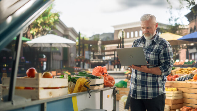 Successful Senior Farmer Working on a Laptop Computer, Writing Emails to Suppliers to Orders Produce for His Organic Street Vendor Stall with Natural Fruits and Vegetables