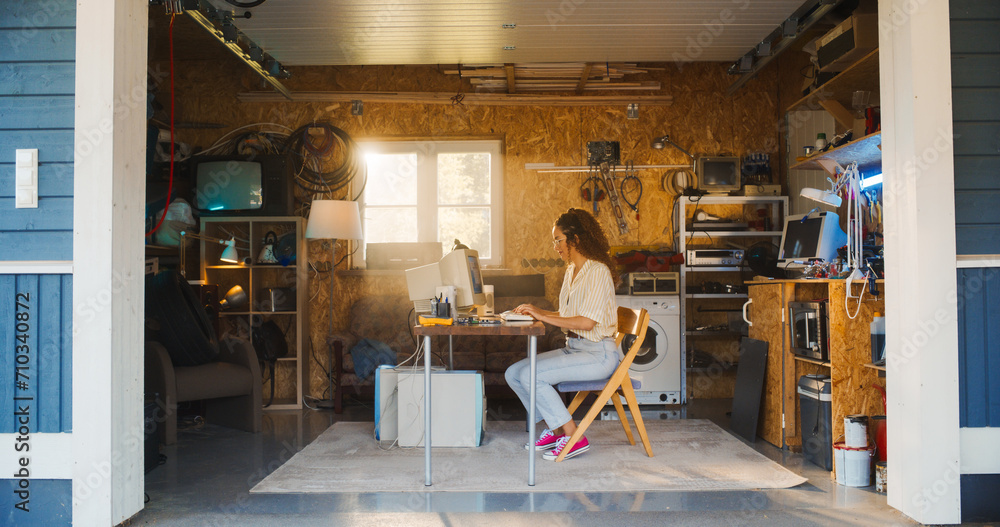 Poster Hispanic Female Software Developer Programming On Old Desktop Computer In Retro Garage With Random Appliances. Woman Working On Innovative Online Service Startup Company In Nineties. Nostalgia Concept
