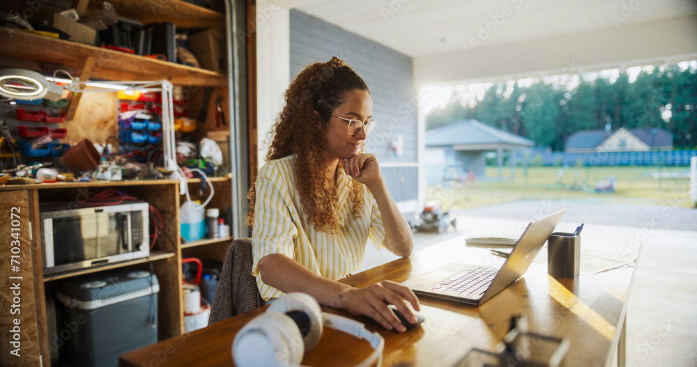 Poster happy hispanic female engineering student sitting at desk and using laptop computer in opened garage