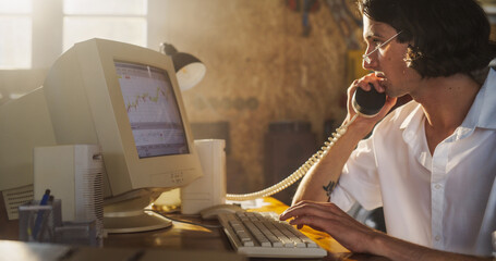 Caucasian Male Trader Talking On Landline Phone And Using Old Desktop Computer In Retro Garage....