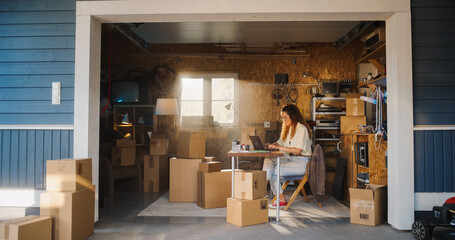 Hispanic Businesswoman Using Laptop Computer In Garage. Female Small Business Owner Filling Orders From Online Customers. Products In Cardboard Boxes Ready For Shipping. Entrepreneur Working At Home. - obrazy, fototapety, plakaty