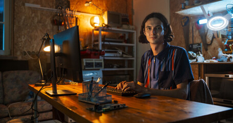 Portrait of Caucasian Male Software Engineer Programming On Desktop Computer In Home Garage, Looking At Camera And Smiling. Ambitious Man Working Innovative Online Service Startup Company at Night.