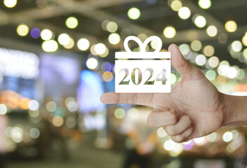 Gift box happy new year 2024 flat icon on finger over blur light and shadow of shopping mall,...