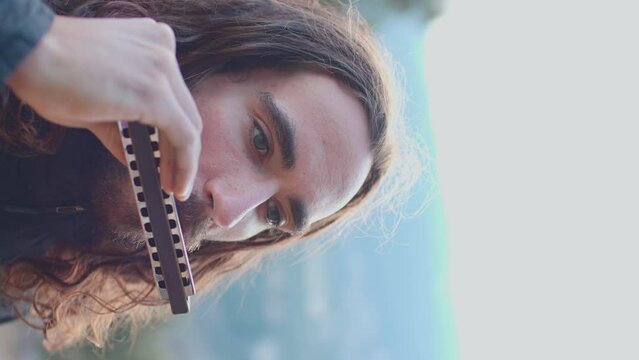 vertical shot. the musician enjoys playing the harmonica while standing on the street. a young man with long hair plays the harmonica.