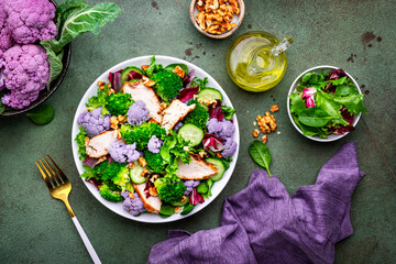 Healthy salad from purple cauliflower, broccoli, chicken breast, fresh cucumbers, onion, radicchio, spinach with walnuts, green table background, top view